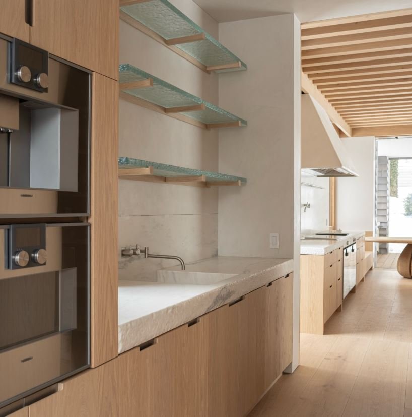 Galley kitchen with honed Danby counters, dark ovens, and glass shelving